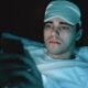 Young man lying in bed wearing a blue sleep mask on his forehead and looking at a smartphone in dim lighting.