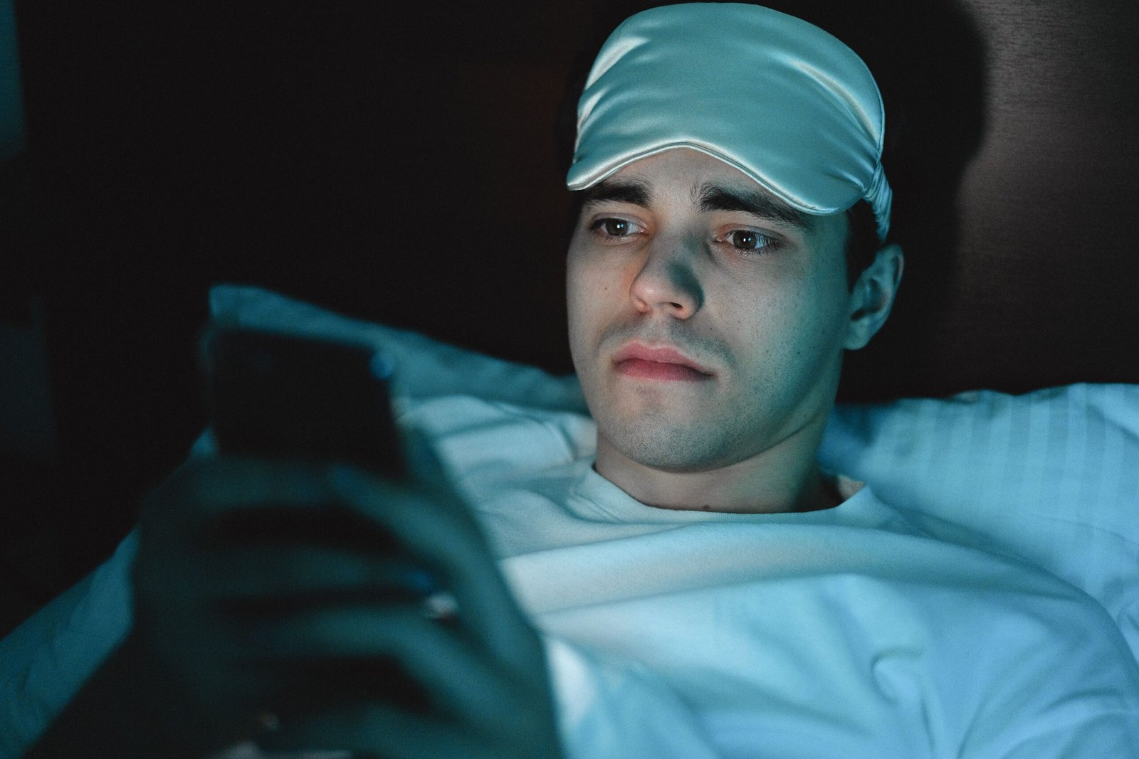 Young man lying in bed wearing a blue sleep mask on his forehead and looking at a smartphone in dim lighting.