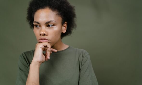 A young woman with natural curly hair resting her chin on her hand and looking thoughtfully to the side.