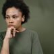 A young woman with natural curly hair resting her chin on her hand and looking thoughtfully to the side.