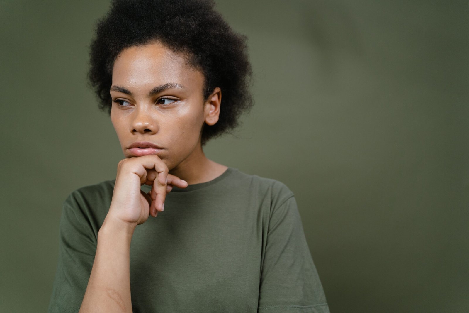 A young woman with natural curly hair resting her chin on her hand and looking thoughtfully to the side.